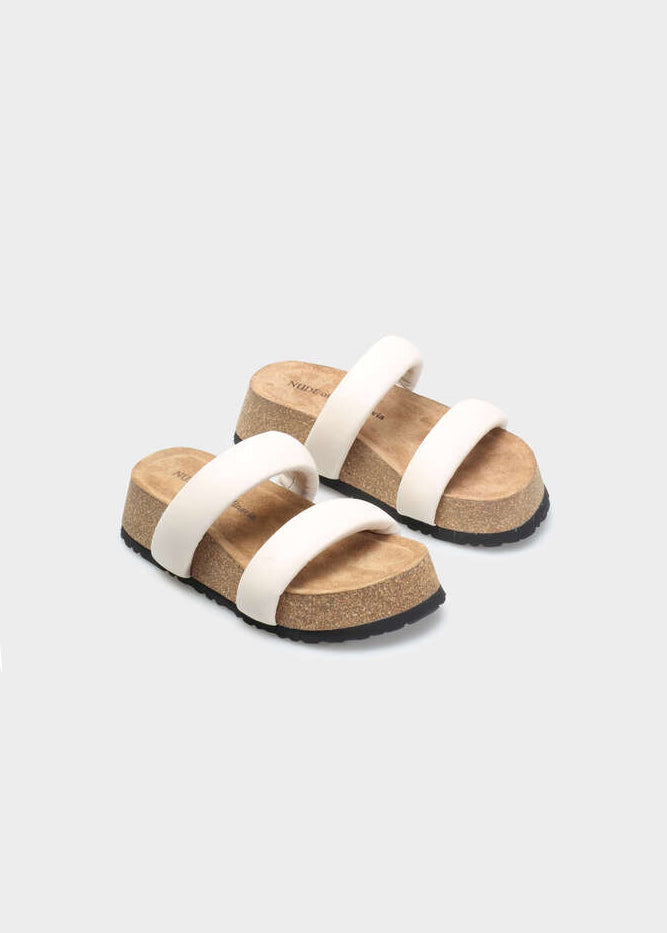 A pair of platform slide sandals with wide off-white straps, cork-look midsoles, and black outsoles, shown on a light gray background.