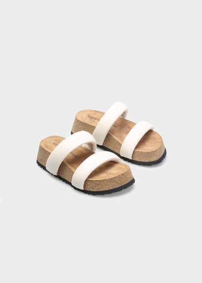 A pair of platform slide sandals with wide off-white straps, cork-look midsoles, and black outsoles, shown on a light gray background.